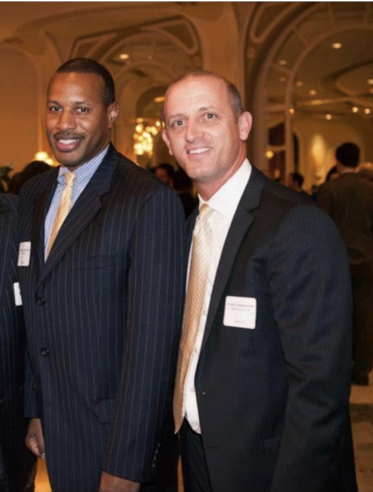 Two men in suits with name badges smiling at a formal networking event